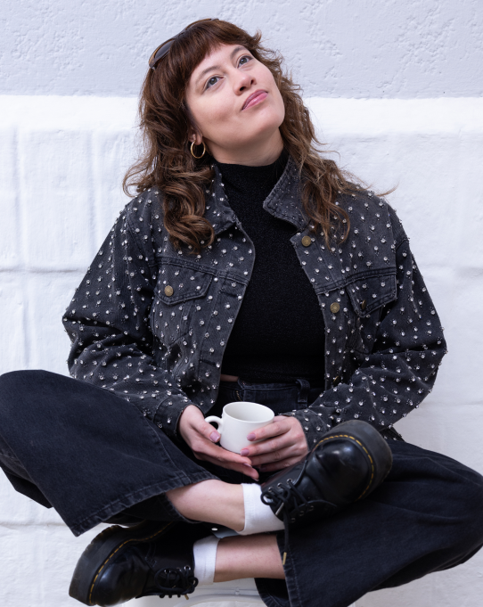 Xiomara sitting cross-legged against a white wall, holding a coffee cup and smiling upward. She has curly brown hair and wears a black polka-dot jacket.